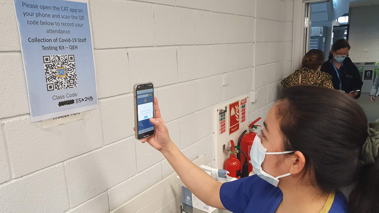 NHS staff member scans a QR code on a poster with the text 'Please open the CAT app on your phone and scan the QR code below to record your attendance. Collection of Covid-19 staff testing kit - QEH. Class Code: 55A24B.’